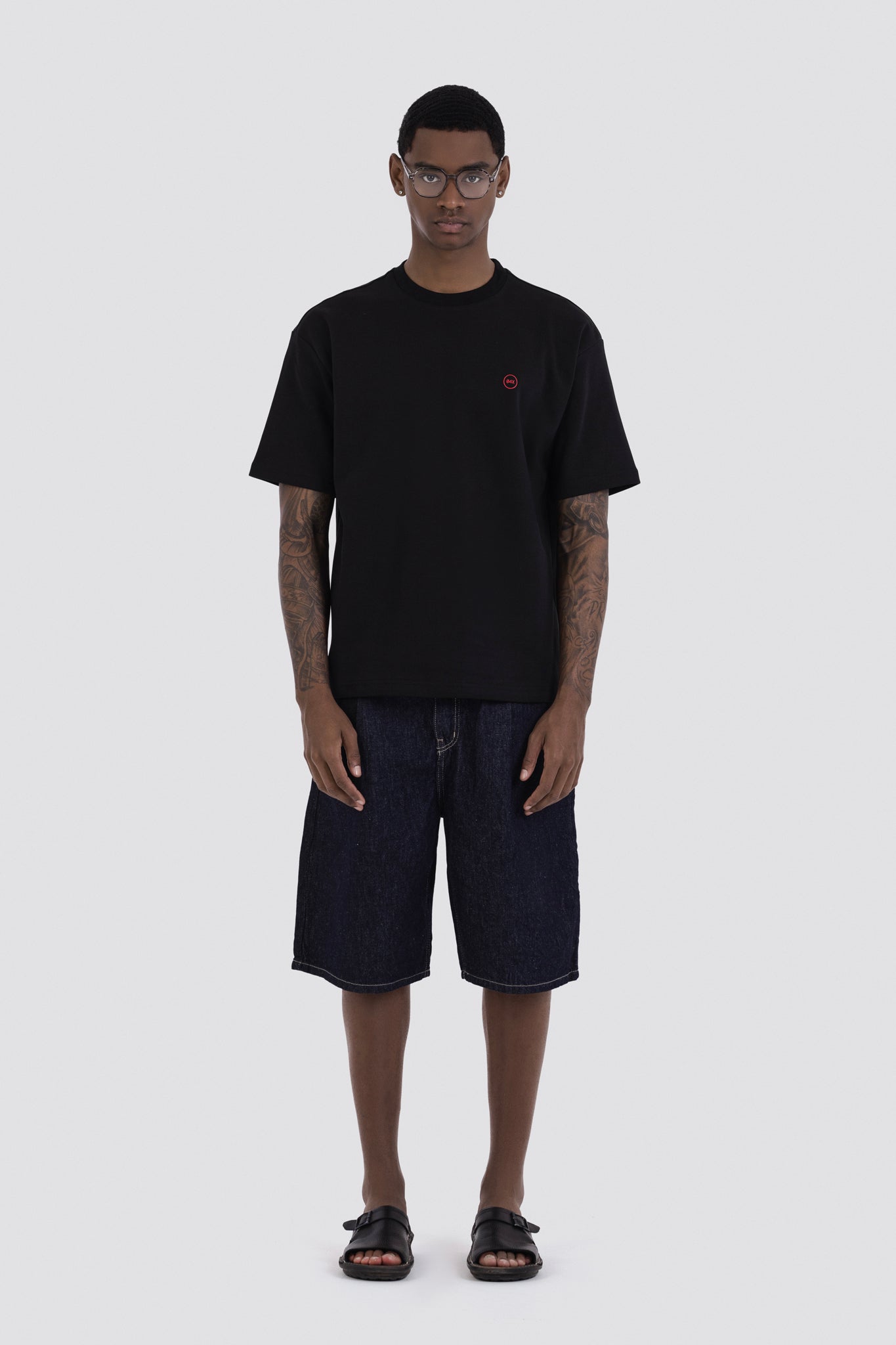 Person wearing a black t-shirt with a small red logo and dark shorts on a white background