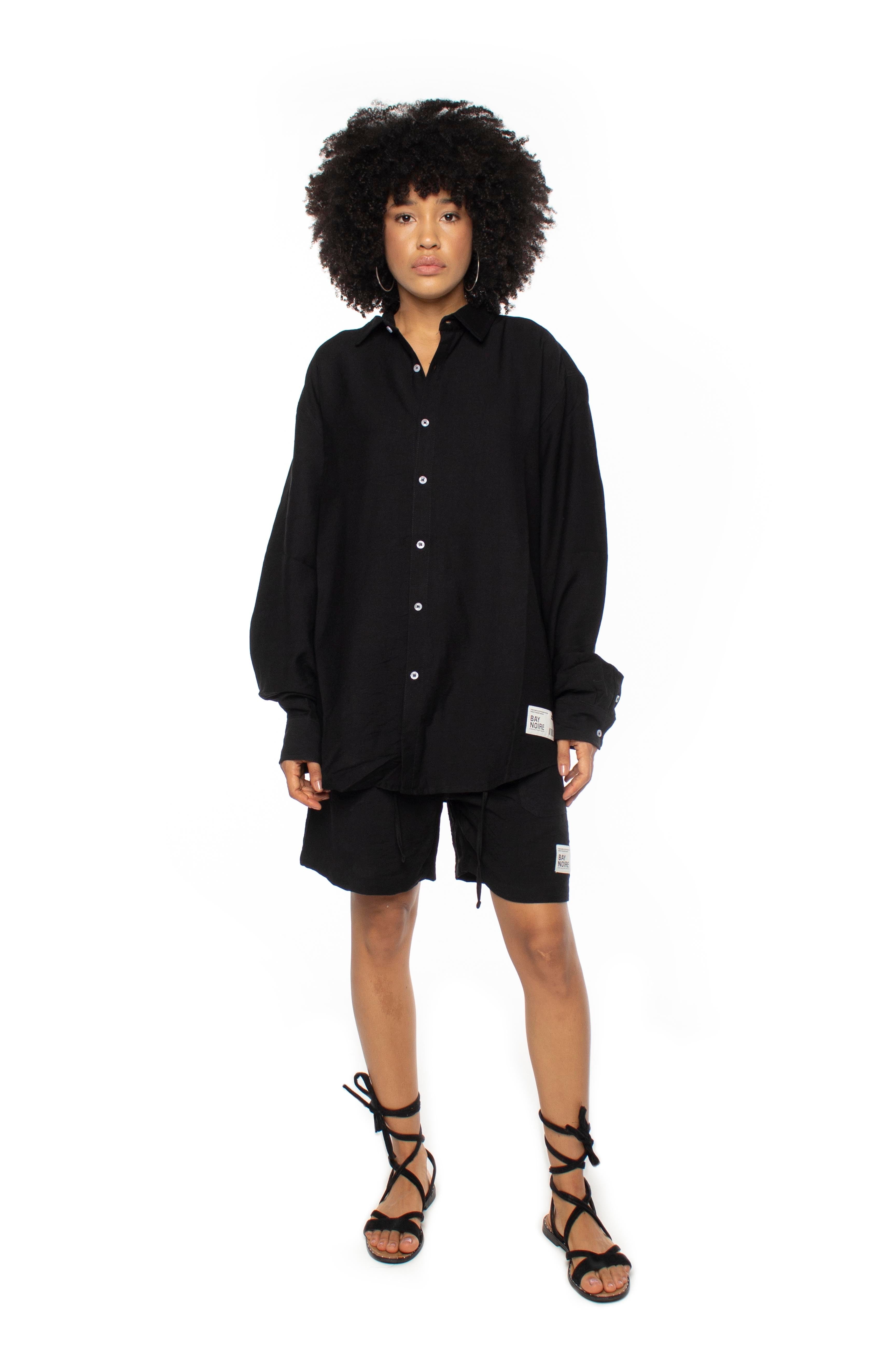 A person wearing a black oversized linen long sleeve shirt with button-up closure and button barrel cuffs. The shirt is oversized and there is a patch on the front.